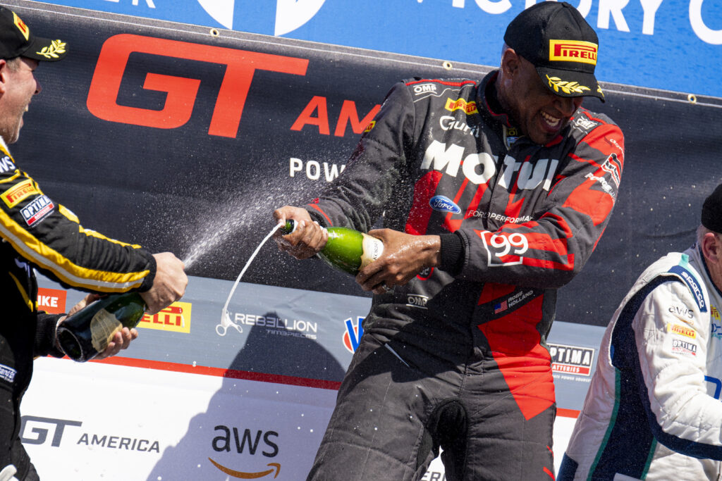 Racecar driver Robb Holland, wearing his driving suit, celebrates by spraying a bottle of champagne. 